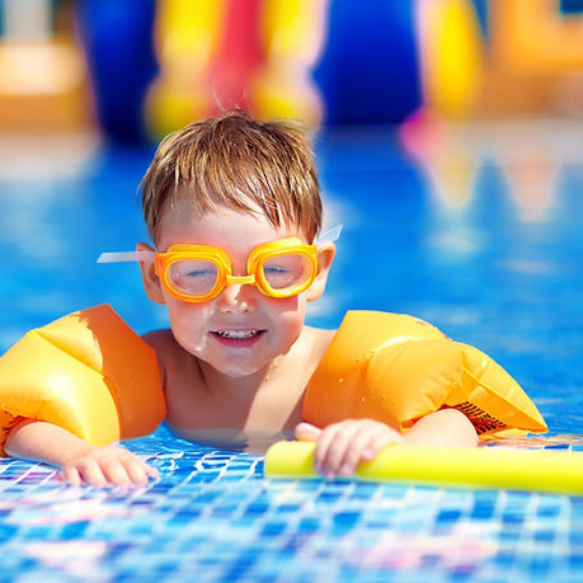 Child in pool wearing orange goggles and arm floaties, holding a yellow pool noodle.