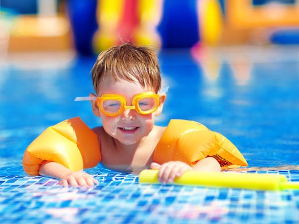 11062b_33d99a10ea964c7e8d2cfaec0dd84c33mv2_ea4b03 Child in pool wearing orange goggles and arm floaties, holding a yellow pool noodle.