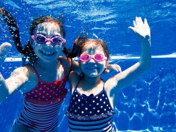Two children in swimsuits and goggles underwater in a pool, smiling and waving.