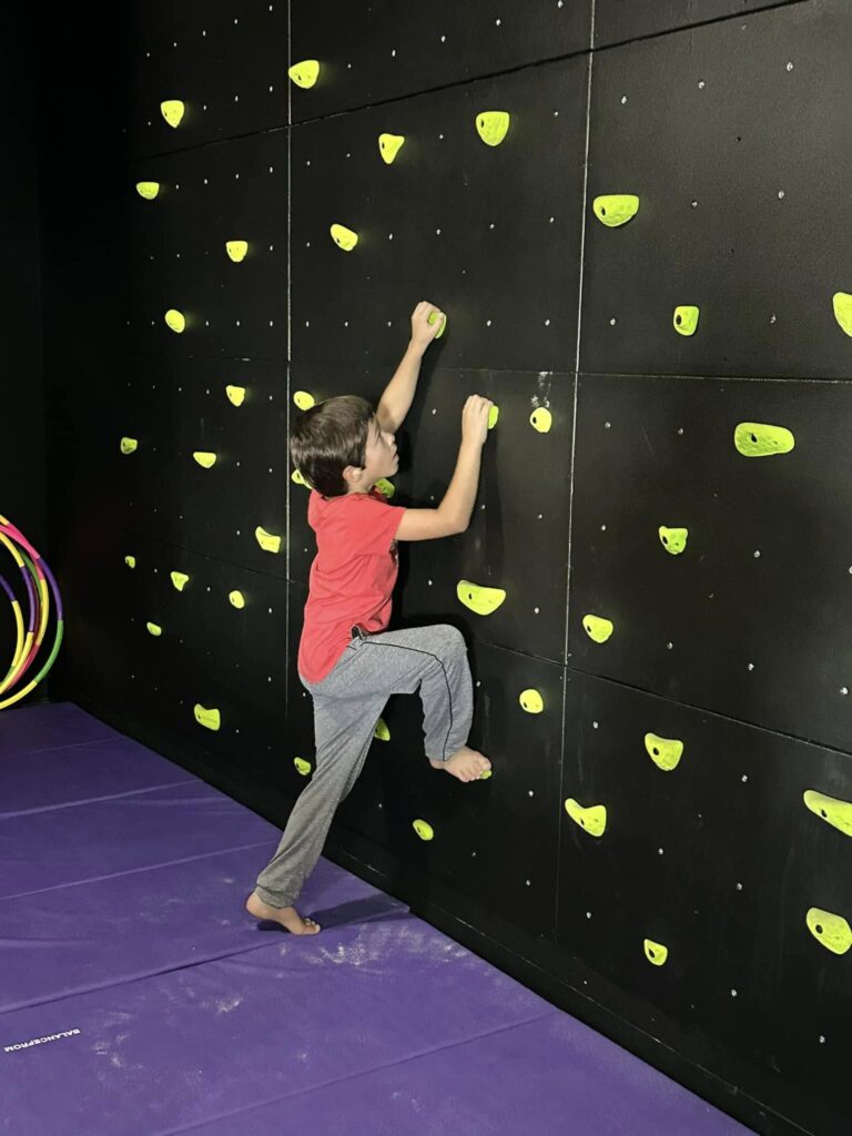 475896630_122182391258119106_6686849138882666119_n-768×1024 Child climbing an indoor bouldering wall with yellow holds on purple mats.