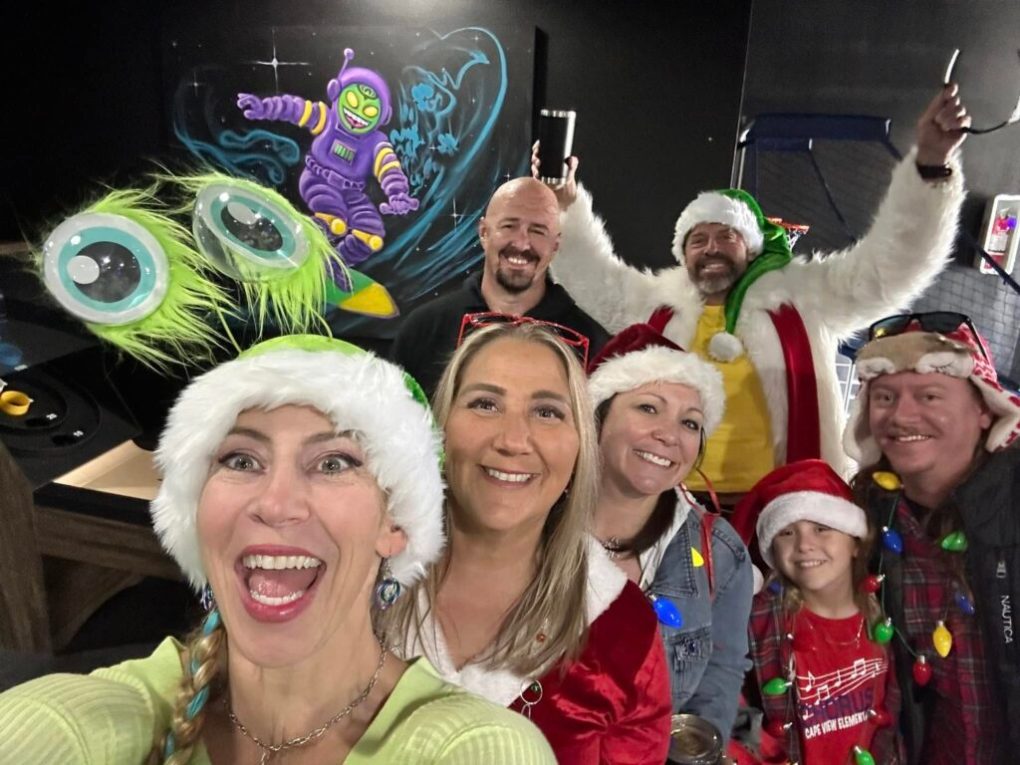 476086422_122182391198119106_7539642171561364854_n-1024×768 Group of people in festive attire with Santa hats and decorations, smiling for a selfie indoors.