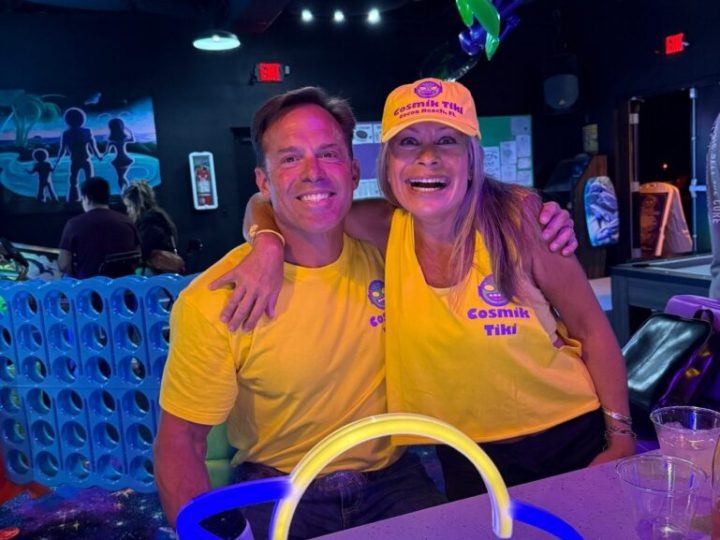 487481686_122190869114119106_1566280720143502342_n-769×1024 Two people in yellow shirts and hats, with neon space-themed decor, smile at a bar with a glowing planet lamp.