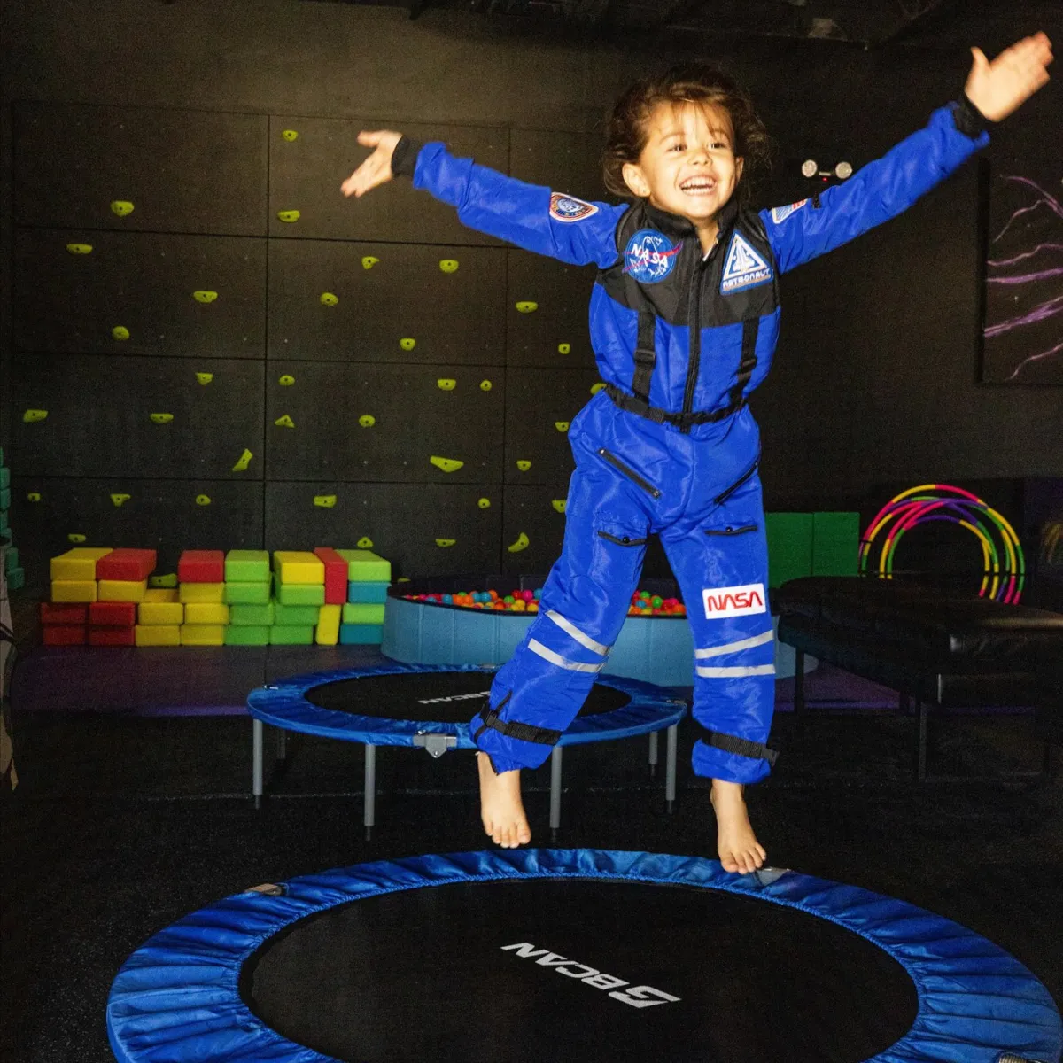 Child in astronaut suit jumping on trampoline indoors with colorful background.