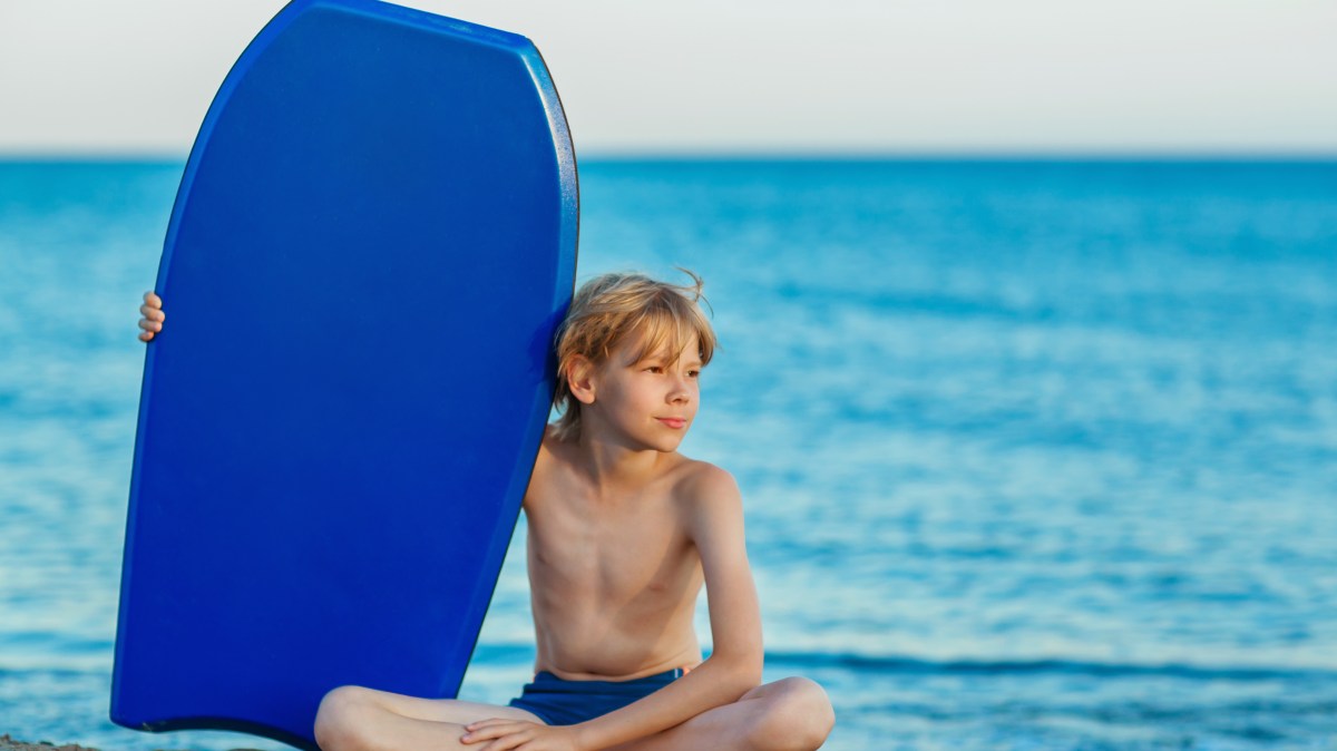 Boy sitting on beach with blue boogie board, ocean in background.