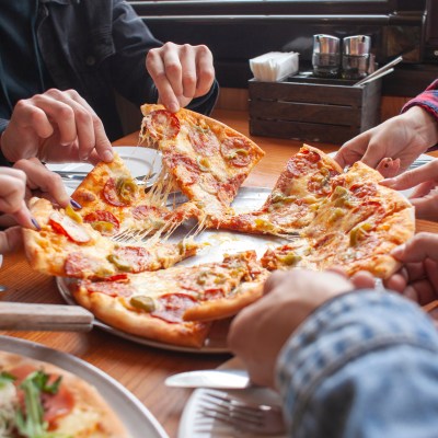 People grabbing slices of pizza from a platter on a wooden table.