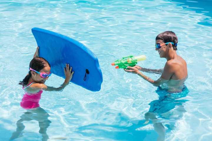 Child with board playing in pool with person holding water gun.