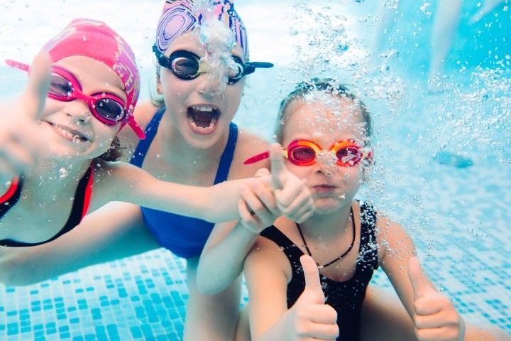 Three kids underwater in a pool, wearing goggles, giving thumbs up.