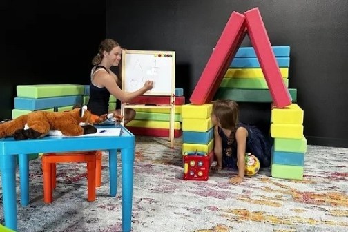 Woman teaching child in playroom with toy blocks and an easel with letters.