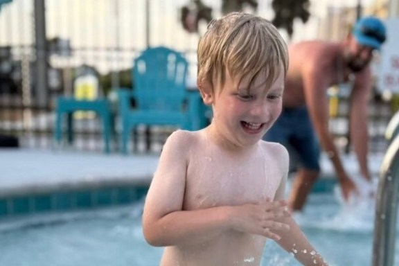 Smiling child splashing in a shallow pool with a man in the background.