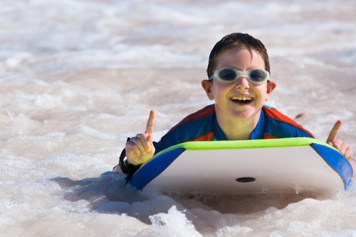 Child with goggles smiling, riding a bodyboard on waves at the beach.