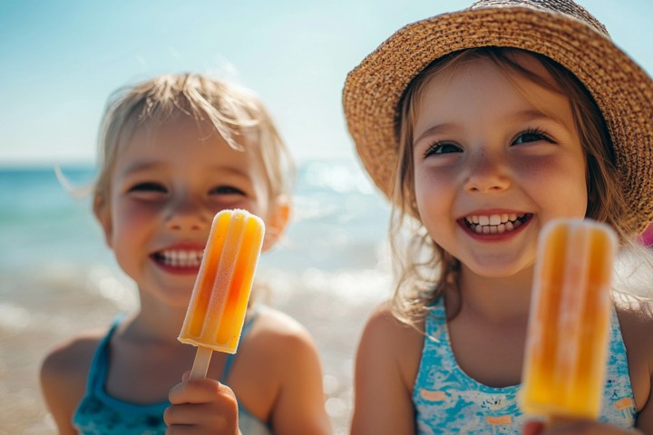 Two smiling children at the beach holding orange popsicles; one wears a hat.