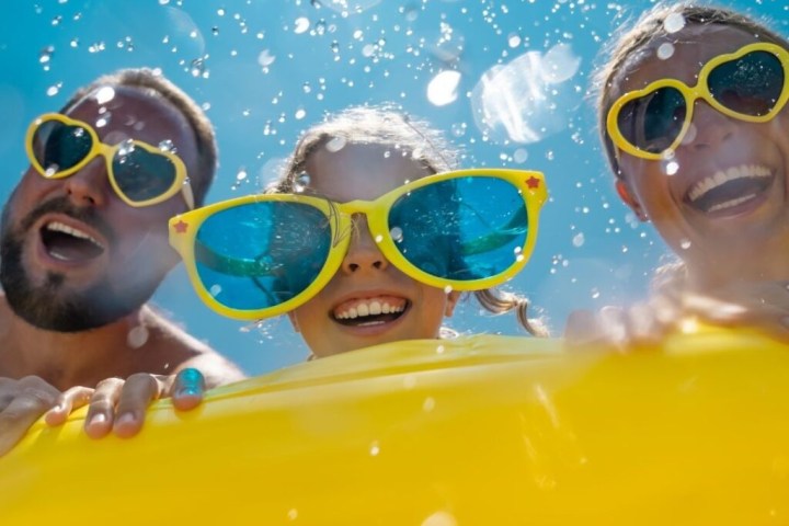 Three people in yellow sunglasses smiling behind a yellow inflatable in a pool with splashing water.