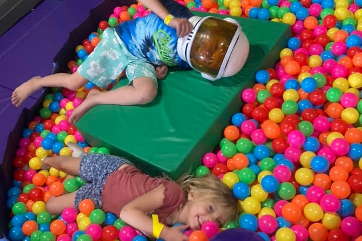 Two kids playing in a colorful ball pit, one wearing an astronaut helmet.