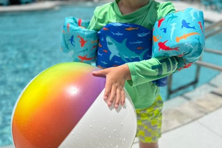 Child in swim gear holding a colorful beach ball by a pool.