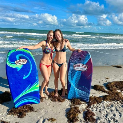 Two people in swimwear holding surfboards on sandy beach with seaweed and ocean in the background.