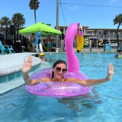 Woman in a pool on a pink flamingo float, smiling with hands raised under a sunny sky.