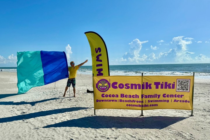 Person on beach with blue flag next to yellow 'Cosmik Tiki' sign under clear sky.
