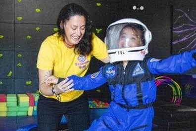 Woman helps child in astronaut suit practicing balance on a play mat in a colorful room.