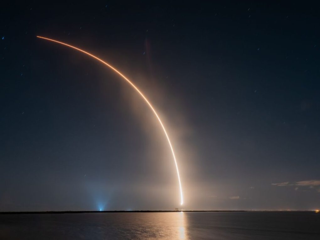 Where-Brevard-Comes-to-Play-19-1024×1024 Long exposure of a rocket launch at night over water, forming an arc of light in the sky.