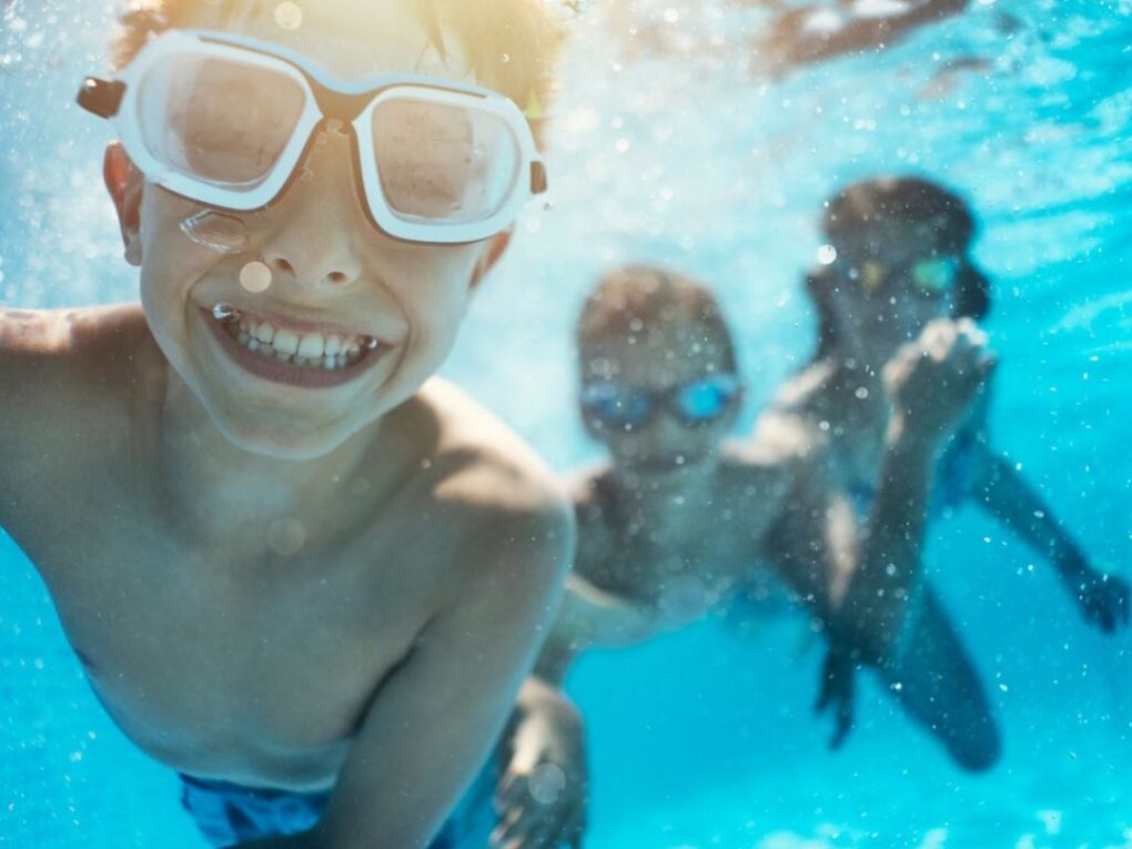 Three children swimming underwater in a pool, wearing goggles and smiling.