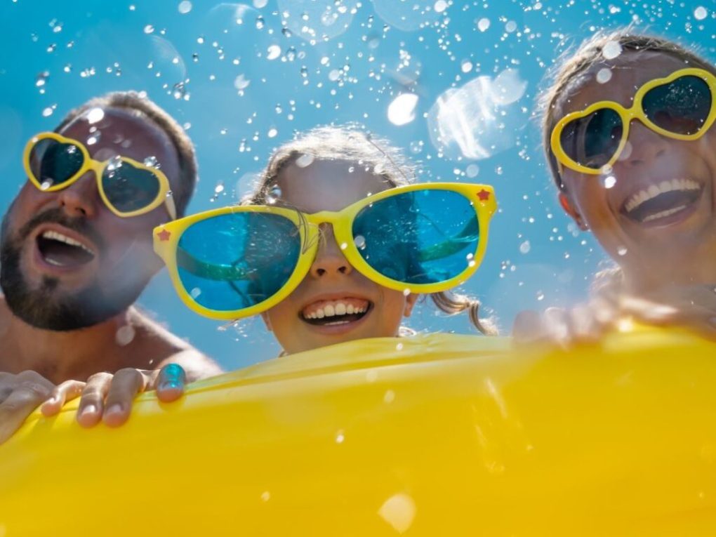 Where-Brevard-Comes-to-Play-31-1024×1024 Three people wearing large yellow sunglasses having fun with water splashes on a sunny day.