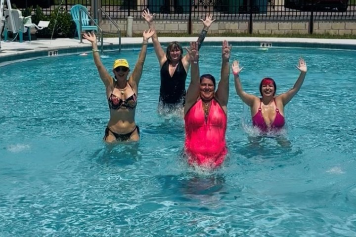 Four people in a pool, raising their hands, smiling in swimsuits.