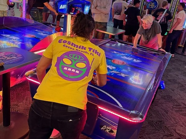 e4c324_6af4f660d6374a55b9bdb5600 Two people playing air hockey in an arcade with colorful lights and other games in the background.