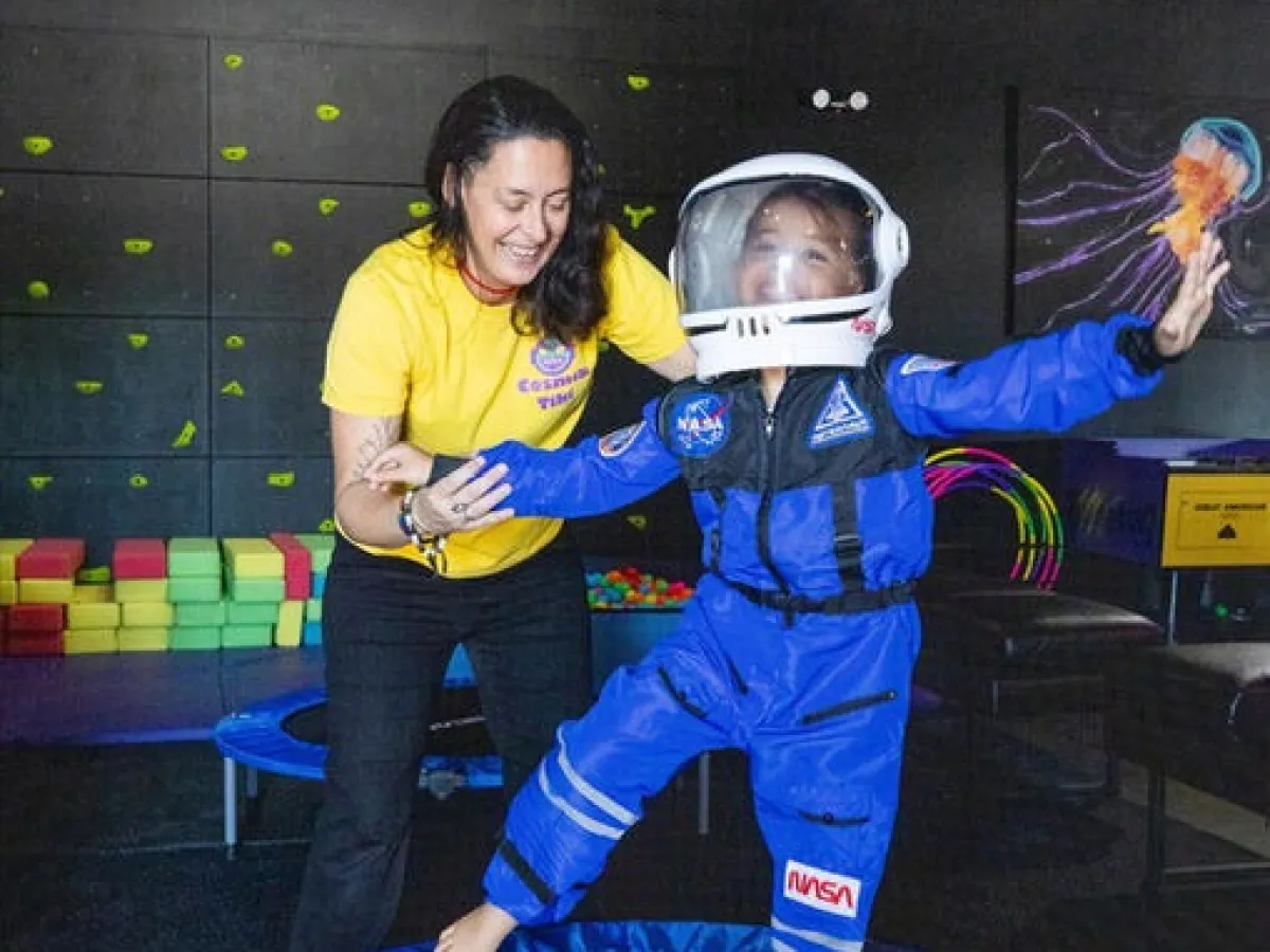 Child in astronaut suit on trampoline assisted by woman in yellow shirt.