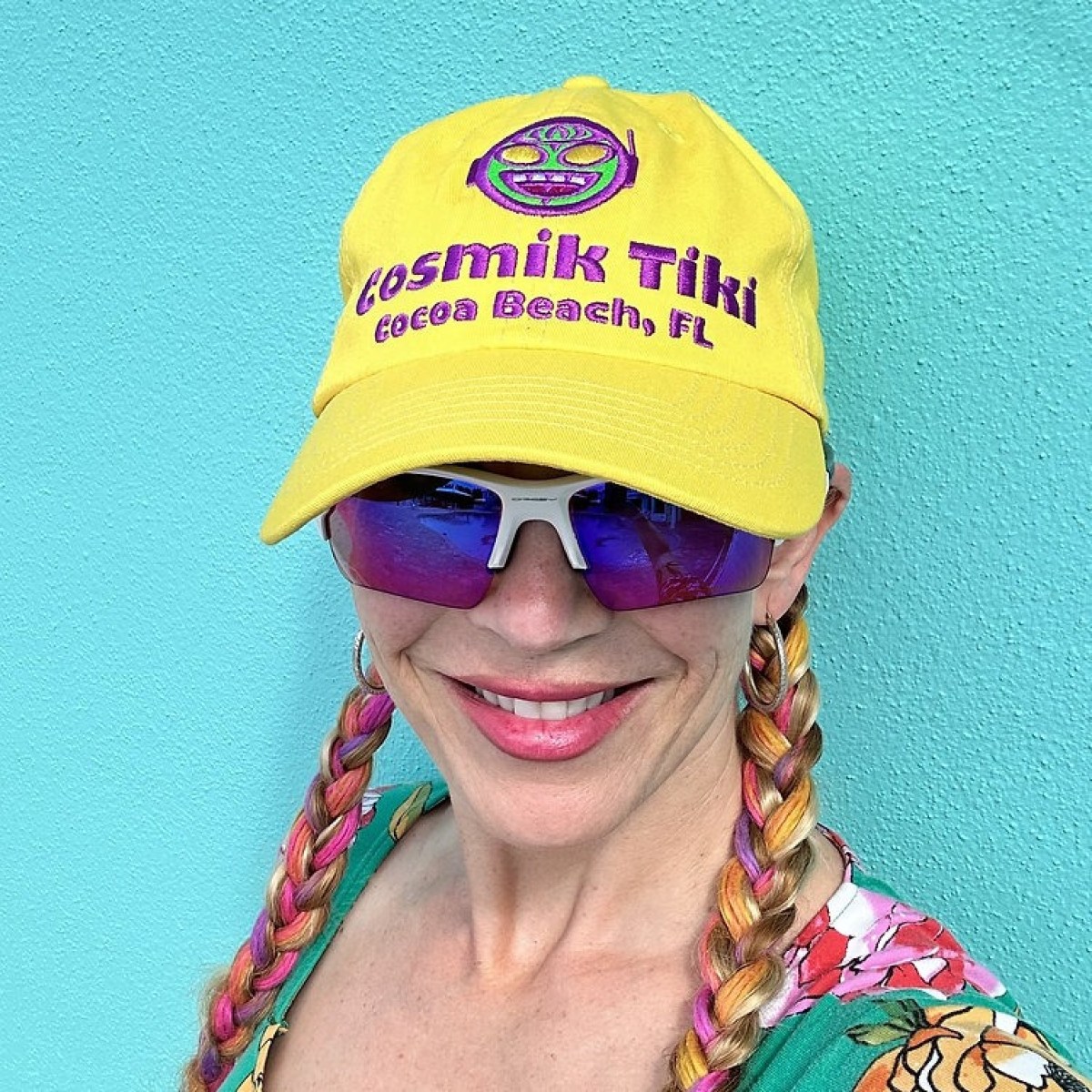 Person wearing yellow hat and sunglasses, smiling in front of a turquoise wall.