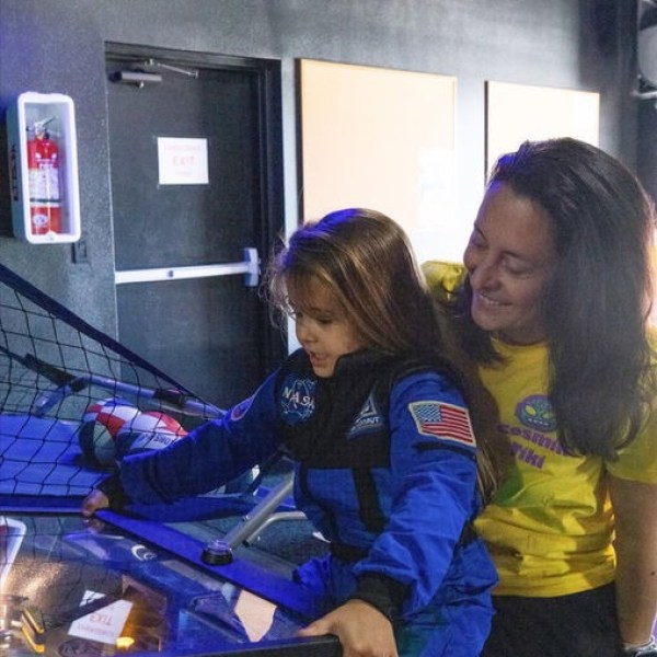 Child in astronaut suit playing pinball, with adult smiling beside them.