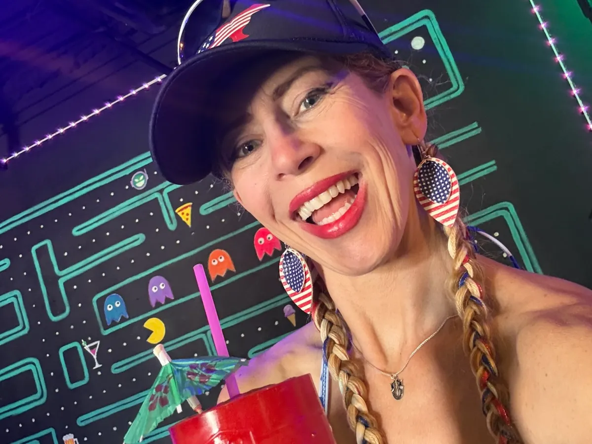 Person with braids and sunglasses holding a red drink at a retro-themed arcade.